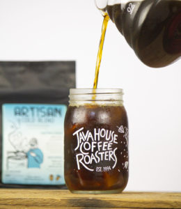 cold brew being poured into a Java House Coffee roaster mason jar, a back of artisan cold blend beans is in the background